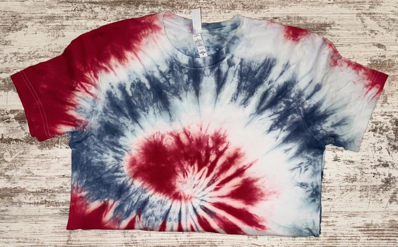 Pode incluir: Uma camiseta de manga curta com um padr&atilde;o tie-dye em vermelho, branco e azul. O design irradia do centro, criando um efeito espiral. A camisa &eacute; feita de um material macio.