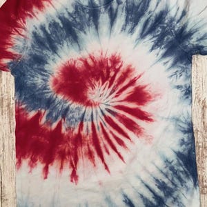 Pode incluir: T-shirt de manga curta com um padr&atilde;o tie-dye em vermelho, branco e azul. O design apresenta um padr&atilde;o em espiral com vermelho no centro, rodeado por tons brancos e azuis. A camisa &eacute; exibida em uma superf&iacute;cie de madeira.