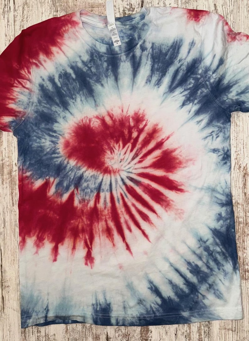 Pode incluir: T-shirt de manga curta com um padr&atilde;o tie-dye em vermelho, branco e azul. O design apresenta um padr&atilde;o em espiral com vermelho no centro, rodeado por tons brancos e azuis. A camisa &eacute; exibida em uma superf&iacute;cie de madeira.