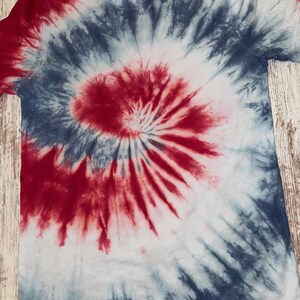 Pode incluir: Uma camiseta branca com um padr&atilde;o espiral tie-dye vermelho, branco e azul. O corante vermelho est&aacute; concentrado no centro, com azul e branco irradiando para fora. A camiseta tem mangas curtas e gola redonda.
