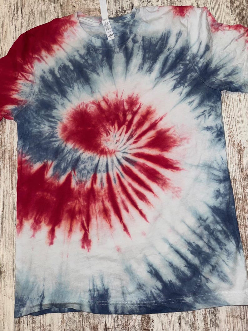 Pode incluir: Uma camiseta branca com um padr&atilde;o espiral tie-dye vermelho, branco e azul. O corante vermelho est&aacute; concentrado no centro, com azul e branco irradiando para fora. A camiseta tem mangas curtas e gola redonda.