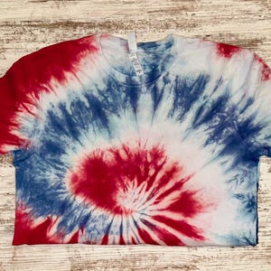 Pode incluir: Uma camiseta tie-dye vermelha, branca e azul. A camiseta de manga curta apresenta um design em espiral, com vermelho no centro, passando para azul e branco. A camiseta est&aacute; colocada em uma superf&iacute;cie de madeira clara.