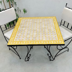 May include: A yellow and white mosaic tile top table with a black wrought iron frame and two matching chairs with white cushions.