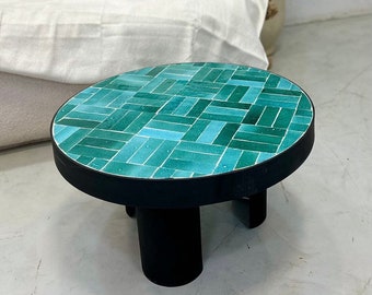 Op de afbeelding: Een ronde salontafel met een tegelblad in tealblauw en een zwarte houten basis met drie poten.