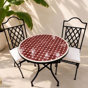 Peut inclure: Un ensemble bistro composé d'une table ronde au plateau en mosaïque rouge foncé et de deux chaises en métal noir avec un dossier en treillis et des coussins d'assise blancs. La table possède un cadre fin en métal noir à quatre pieds.