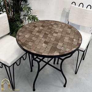 Peut inclure: Une table de bistrot ronde avec un plateau en mosaïque de carreaux bruns en forme d'écailles et un cadre en métal noir, accompagnée de deux chaises en métal noir assorties avec des coussins d'assise blancs. Le plateau présente un motif.