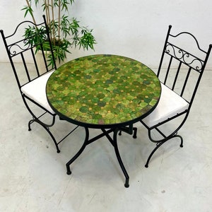 May include: A round green mosaic table with a black wrought iron frame and two matching chairs with white cushions. The table and chairs are perfect for a small patio or balcony.