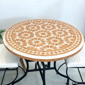 Op de afbeelding: Een ronde tafel met een wit en oranje mozaïek tegelblad en een zwart metalen frame. Twee stoelen met witte kussens en zwarte metalen frames staan aan weerszijden van de tafel.