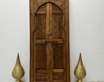 Puede incluir: Una puerta de madera grande y tallada con dos paneles. La puerta es marrón y tiene un diseño tradicional marroquí. La puerta está enmarcada por un marco de madera.