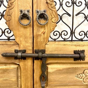 May include: A pair of wooden doors with intricate carvings and a metal latch. The doors are painted a light brown color and have a rustic look. The metal latch is black and has a simple design.