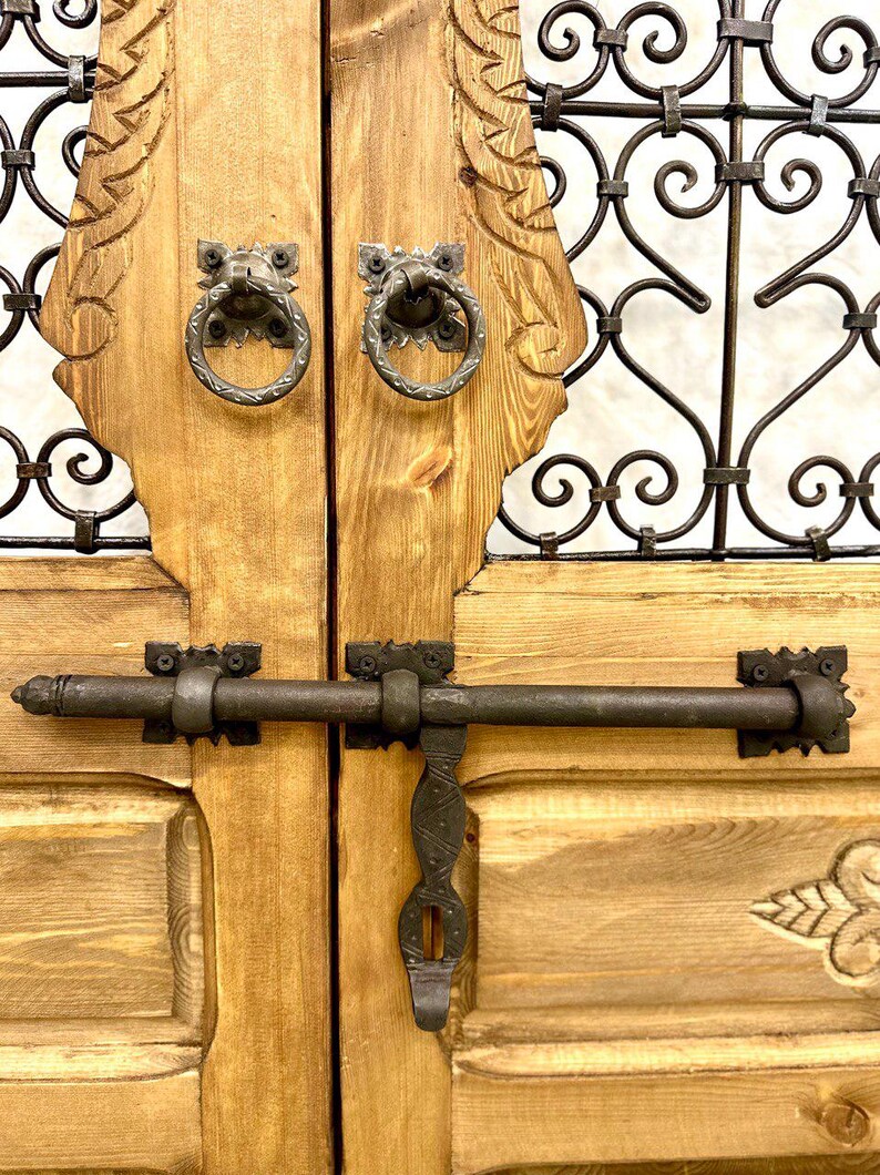 May include: A pair of wooden doors with intricate carvings and a metal latch. The doors are painted a light brown color and have a rustic look. The metal latch is black and has a simple design.