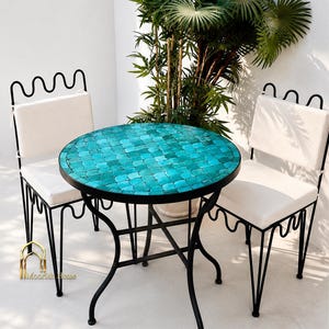 May include: A round bistro table with a turquoise fish-scale mosaic top and a black metal frame, accompanied by two matching black metal chairs featuring white seat cushions and a wavy top rail design, with the text Moorish Home visible.