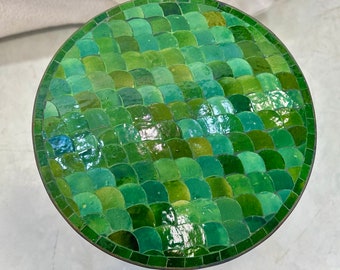 Op de afbeelding: Een ronde mozaïektafel met een groen, schubachtig patroon. Het tafeloppervlak heeft verschillende tinten groene tegels, waardoor een textuur ontstaat. De tafel heeft een donkerbruine metalen rand. De diameter is ongeveer 40 cm.