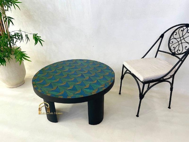 May include: A round, black wooden table with a geometric pattern of green, blue, and gold triangles. The table has four legs and is sitting on a white surface. A black metal chair with a white cushion is in the background.
