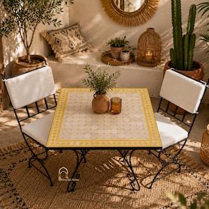Op de afbeelding: Bistroset voor buiten met een vierkante tafel en twee stoelen. De tafel heeft een geel en wit tegelblad en zwarte metalen poten. De stoelen hebben witte kussens en zwarte metalen frames.