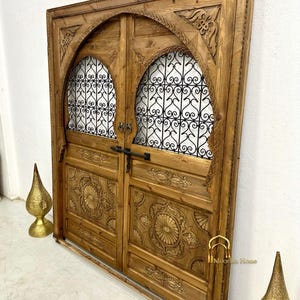 May include: A pair of intricately carved wooden doors with arched tops and metal grilles. The doors are a rich brown color and have a rustic, antique look. The doors are framed by a wooden surround.