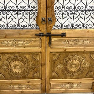 May include: A pair of wooden doors with intricate carved details and a metal latch. The doors have a decorative iron grate design at the top.