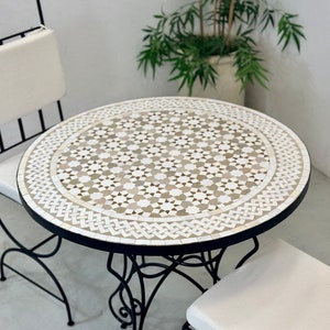 Può includere: Un tavolo rotondo in mosaico di piastrelle bianche e beige con una cornice in metallo nero e due sedie bianche imbottite. Il tavolo ha un motivo geometrico.