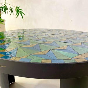 May include: A round mosaic table with a black base and a top made of green, blue, and beige tiles. The table is made of geometric shapes and has a glossy finish.