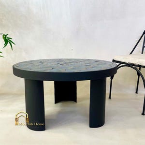 May include: A round black mosaic tile coffee table with three legs. The table is made of black painted wood with a mosaic tile top. The table is sitting on a light gray surface.