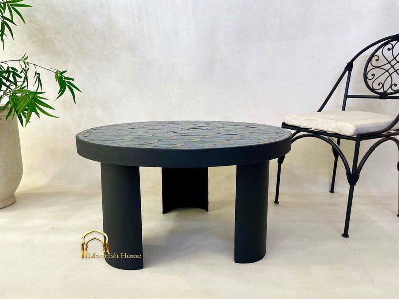May include: A round black mosaic tile coffee table with three legs. The table is made of black painted wood with a mosaic tile top. The table is sitting on a light gray surface.