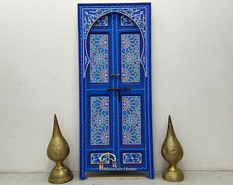 Op de afbeelding: Een blauwe houten deur met ingewikkelde bloemmotieven en een Moorish Home logo. De deur is geschilderd in een levendig blauw met witte en gele accenten. De deur wordt omlijst door een witte muur en twee gouden decoratieve accenten.