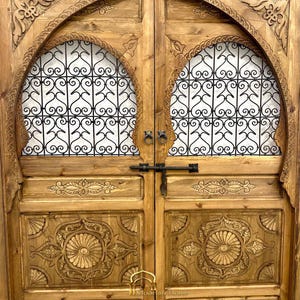 May include: A pair of intricately carved wooden doors with arched tops and metal latticework panels. The doors are painted in a warm brown color and feature decorative floral motifs.