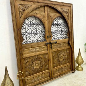 May include: A pair of intricately carved wooden doors with arched tops and metal grilles. The doors are a rich brown color and have a distressed finish. The doors are set against a white wall.