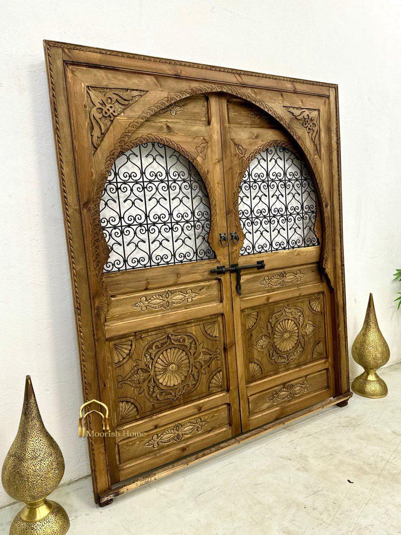 May include: A pair of intricately carved wooden doors with arched tops and metal grilles. The doors are a rich brown color and have a distressed finish. The doors are set against a white wall.