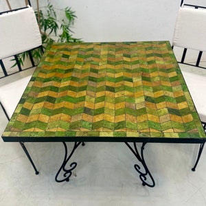Può includere: Un tavolo quadrato con un piano in mosaico di piastrelle verdi e marroni e gambe in metallo nero con un design a scorrimento decorativo.