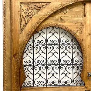 May include: A wooden door with intricate carvings and a metal grate design. The door is painted in a light brown color and has a traditional Moroccan style.
