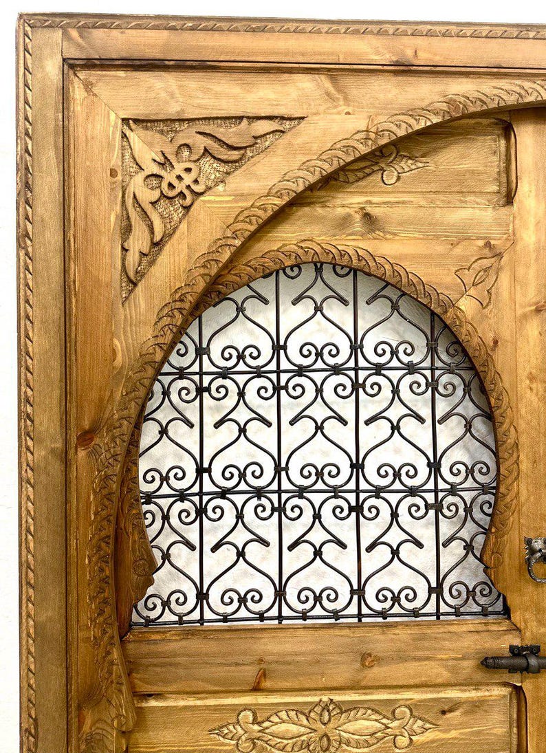 May include: A wooden door with intricate carvings and a metal grate design. The door is painted in a light brown color and has a traditional Moroccan style.
