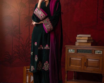 May include: A dark green velvet outfit with pink and gold floral embroidery. The ensemble includes a tunic and trousers, complemented by a magenta scarf. The sleeves and hem are adorned with detailed patterns. The model is wearing magenta heels. The outfit is suitable for special occasions.