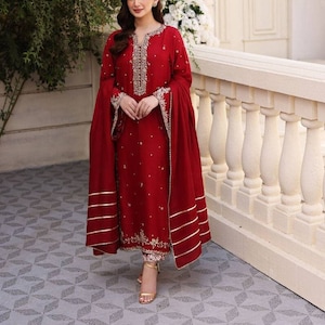 K&ouml;nnte beinhalten: Ein rotes, lang&auml;rmliges Kleid mit goldener Stickerei und einem passenden roten Dupatta. Das Kleid hat einen hohen Ausschnitt und einen langen Rock. Der Dupatta ist &uuml;ber die Schultern der Frau gelegt und hat einen goldenen Rand.
