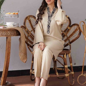 May include: A beige two-piece outfit featuring a long-sleeved tunic with black embroidery around the neckline and cuffs, paired with matching straight-leg pants. The outfit is worn with black heels. The model is seated on a wicker chair.