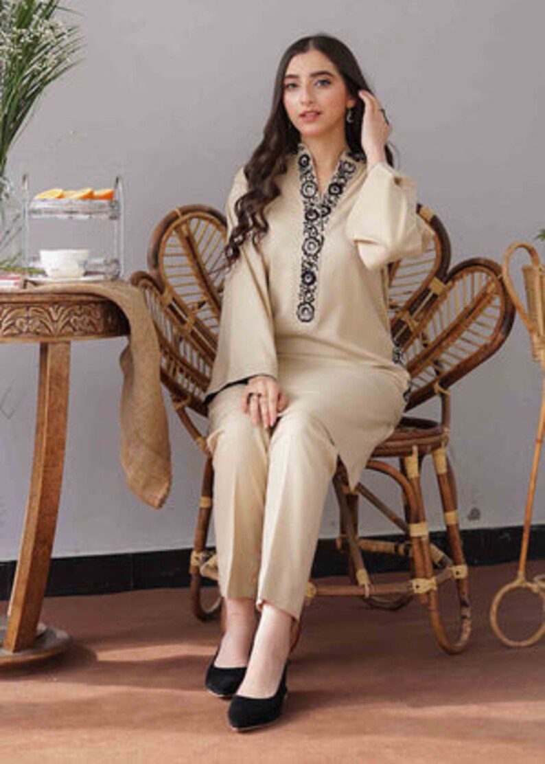 May include: A beige two-piece outfit featuring a long-sleeved tunic with black embroidery around the neckline and cuffs, paired with matching straight-leg pants. The outfit is worn with black heels. The model is seated on a wicker chair.