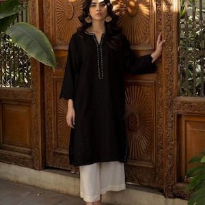 Puede incluir: Una túnica negra de manga larga con cuello en V y ribetes blancos. La túnica se combina con pantalones blancos de pierna ancha y zapatos marrones con bordados florales.