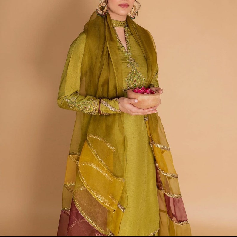 May include: A green embroidered kurta with a matching dupatta. The dupatta has gold and maroon trim and is draped over the shoulders. The kurta has a high neckline and long sleeves. The woman is holding a small bowl of rose petals.