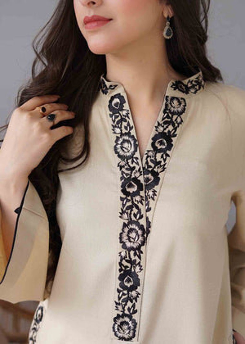 May include: Cream-colored tunic with black floral embroidery along the neckline. The long-sleeved top has a V-neck design and a relaxed fit. The sleeves have a flared design with black trim.