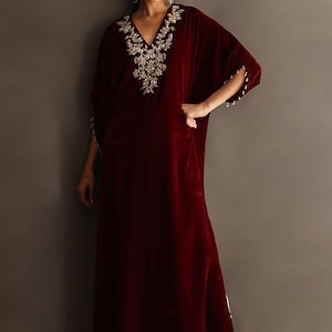 Könnte beinhalten: Langes, bordeauxfarbenes Samtkleid mit V-Ausschnitt und Verzierungen am Ausschnitt. Die Ärmel haben Perlenakzente. Das bodenlange Kleid hat einen Seitenschlitz. Das Model trägt goldfarbene Ohrringe und Schuhe.