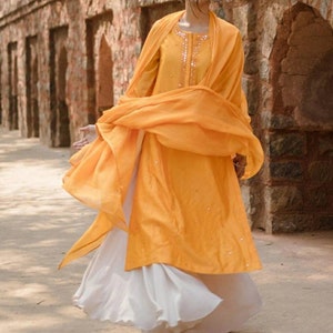 May include: A woman wearing a yellow embroidered kurta with a white skirt and a matching yellow dupatta. The outfit is decorated with small, sparkly embellishments.