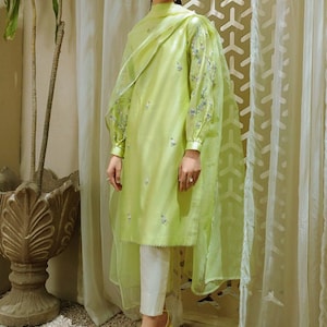 May include: A light green long-sleeved tunic with delicate embroidery and a matching sheer dupatta. The tunic has a high neck and a straight silhouette. The dupatta is draped over the shoulders and has a scalloped edge. The model is wearing white pants and yellow heels.