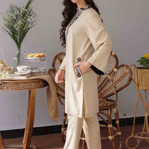 May include: A beige two-piece outfit featuring a long-sleeved tunic and matching pants. The tunic has black trim on the cuffs and decorative embroidery around the neckline and on the pocket. The outfit is worn with black flats.