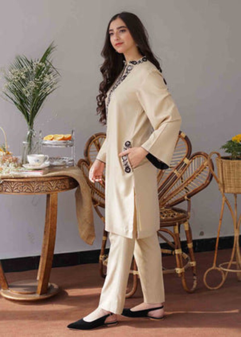 May include: A beige two-piece outfit featuring a long-sleeved tunic and matching pants. The tunic has black trim on the cuffs and decorative embroidery around the neckline and on the pocket. The outfit is worn with black flats.