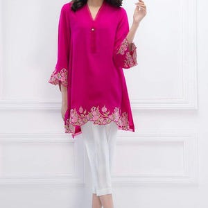 Puede incluir: Una túnica de color rosa fucsia con cuello en V y detalles florales bordados alrededor del dobladillo y las mangas. La túnica se combina con pantalones blancos.