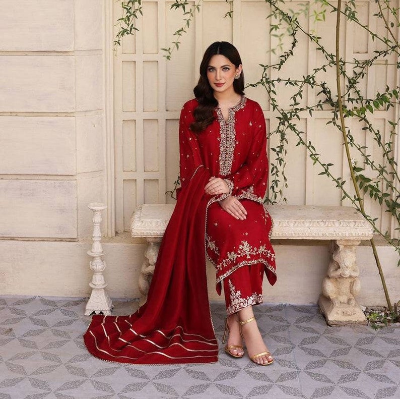 K&ouml;nnte beinhalten: Eine Frau tr&auml;gt ein rotes besticktes Kleid mit einem passenden roten Dupatta. Das Kleid ist mit goldenen Stickereien verziert und das Dupatta hat einen goldenen Saum. Sie sitzt auf einer wei&szlig;en Bank vor einer wei&szlig;en Wand mit gr&uuml;nen Ranken.