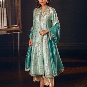 May include: A teal blue embroidered kurta with a matching dupatta. The kurta has a long, flowing silhouette and is adorned with intricate floral embroidery. The dupatta is sheer and has a delicate border. The model is wearing gold trousers and matching heels.
