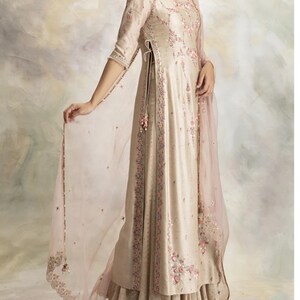 Pu&ograve; includere: Un lungo abito color crema con ricami floreali e un dupatta rosa trasparente. L'abito ha uno spacco laterale e una gonna a strati.