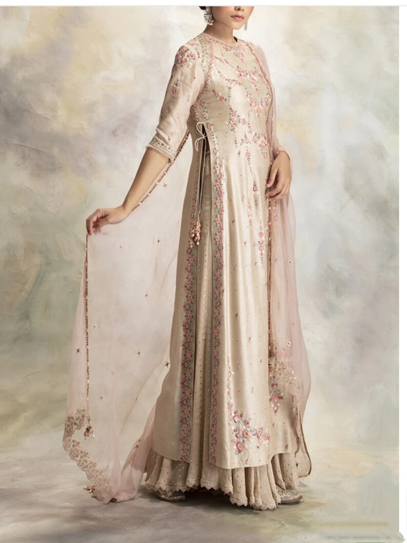 Pu&ograve; includere: Un lungo abito color crema con ricami floreali e un dupatta rosa trasparente. L'abito ha uno spacco laterale e una gonna a strati.