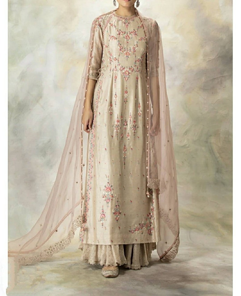 Pu&ograve; includere: Un lungo abito beige con ricami floreali e un dupatta rosa trasparente abbinato. L'abito ha una scollatura rotonda e maniche a tre quarti. Il dupatta ha un bordo in pizzo.